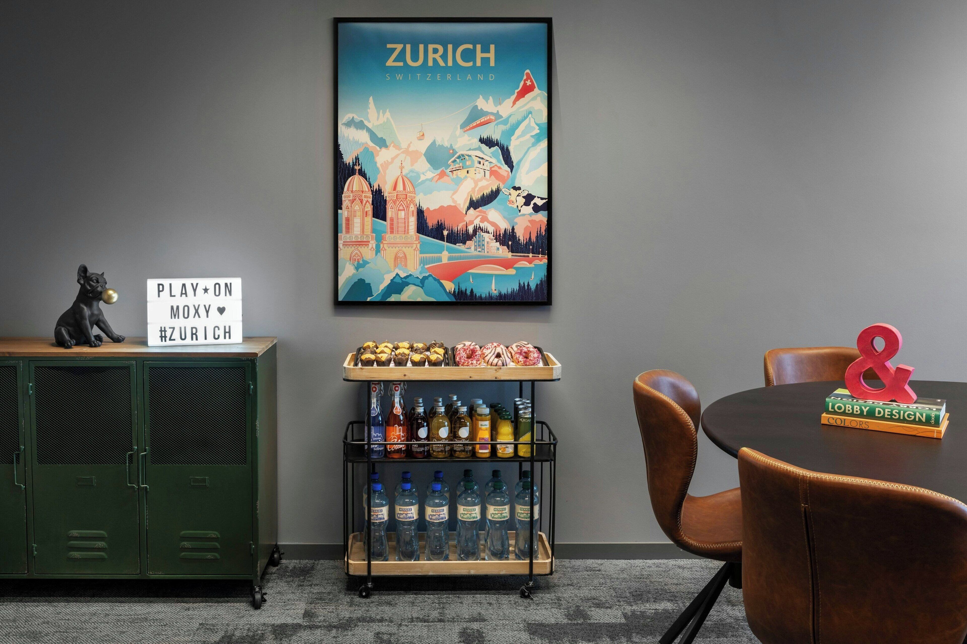 Een kamer met een poster van Zürich, een kast met flessen, een bordje met de tekst PLAY*ON MOXY #ZURICH, en een tafel met een boek en een rood uitroepteken.