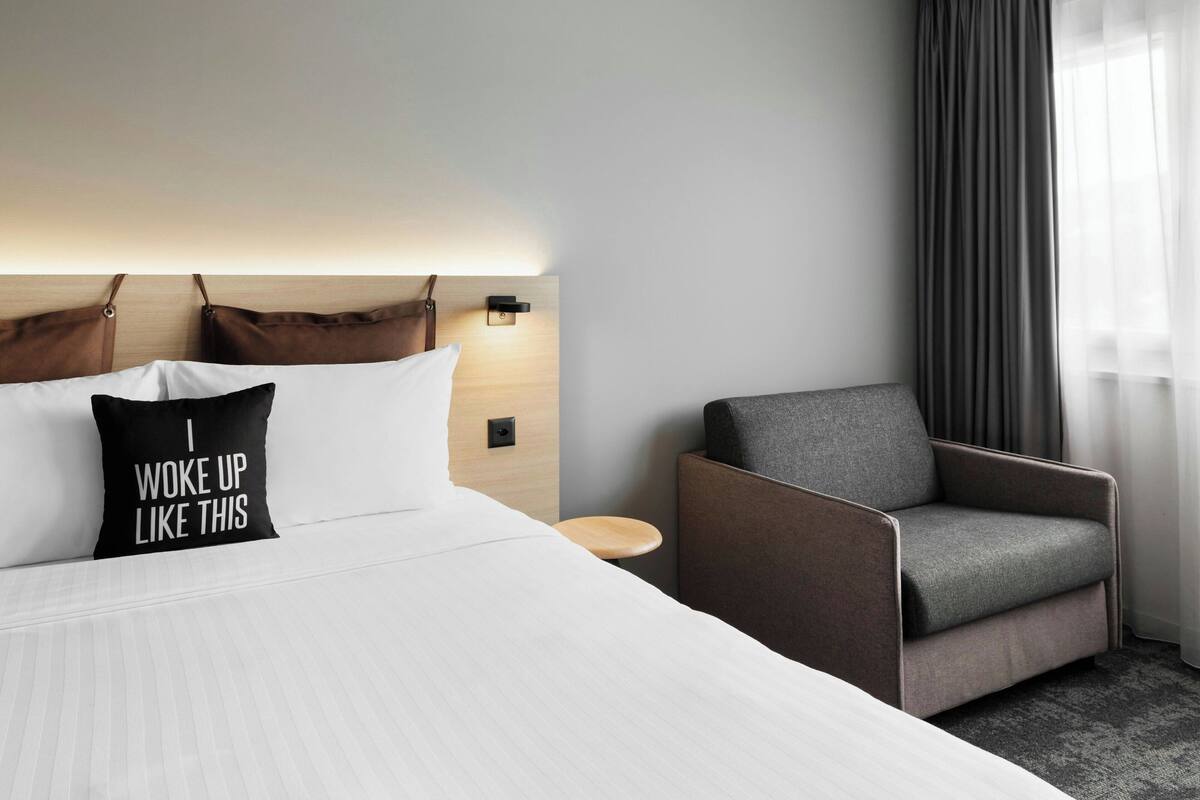 A hotel room with a bed, a chair, a nightstand, and a wall-mounted lamp.