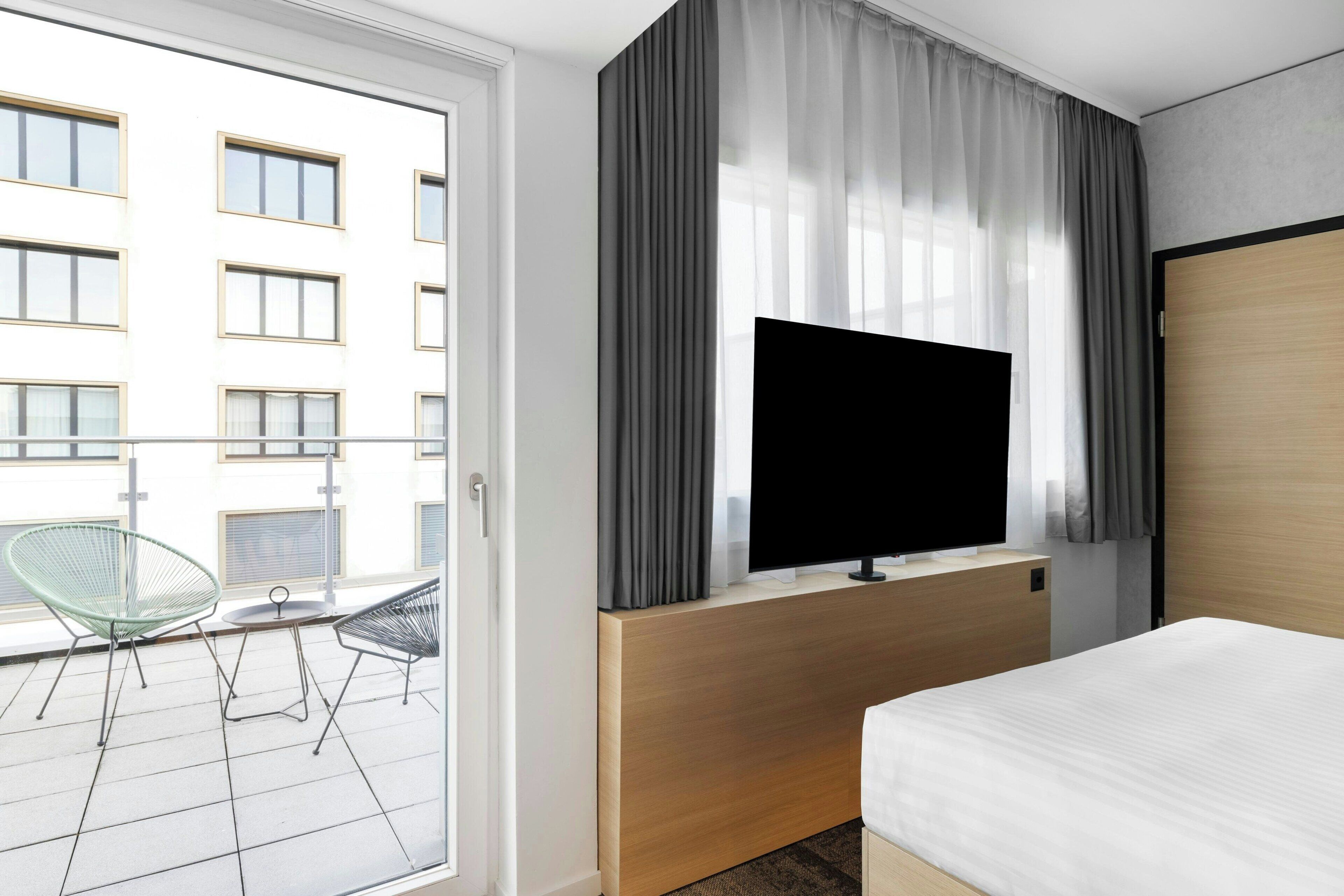 A hotel room with a bed, a TV mounted on a wooden panel, a balcony with a chair and a small table, and a view of a building with multiple windows.