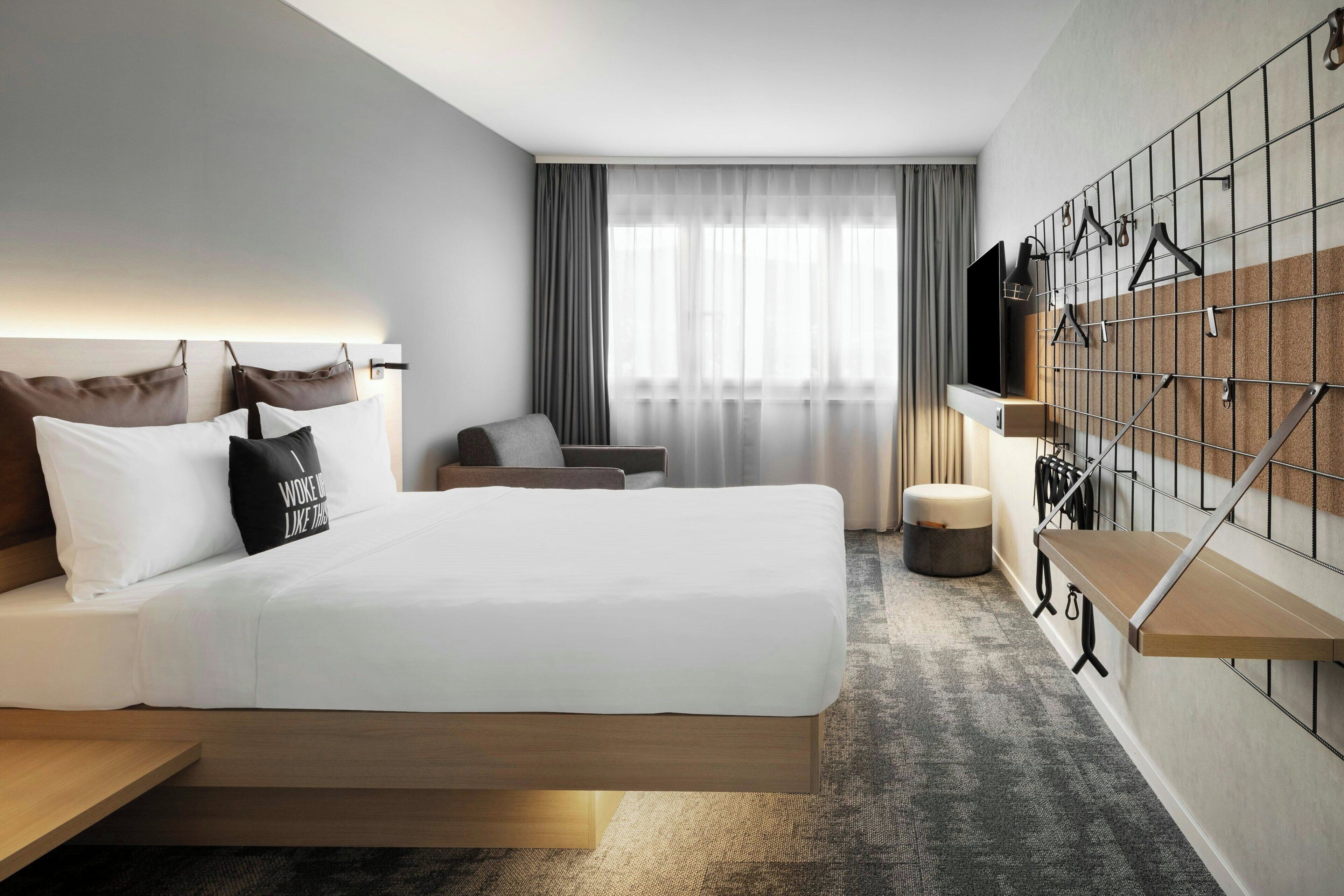A modern hotel room with a large bed, a seating area, a TV, and a wall-mounted storage unit.