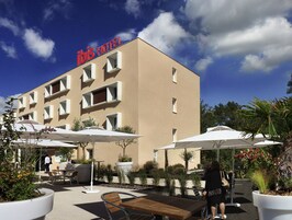 Un espacio exterior con un edificio que dice 'ibis HOTEL', con mesas y sillas afuera y sombrillas.