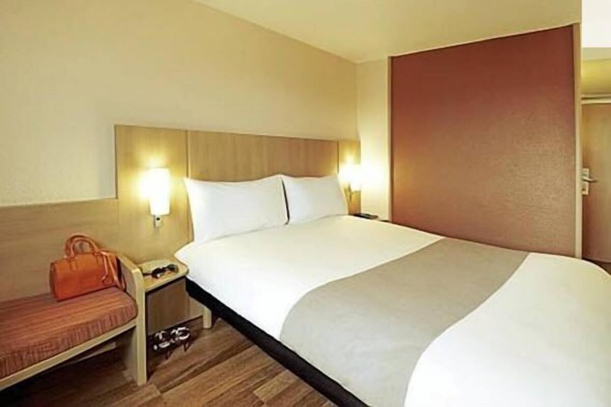 Habitación de hotel con una cama grande, una mesita de noche con lámpara y un pequeño rincón de estar con un bolso.