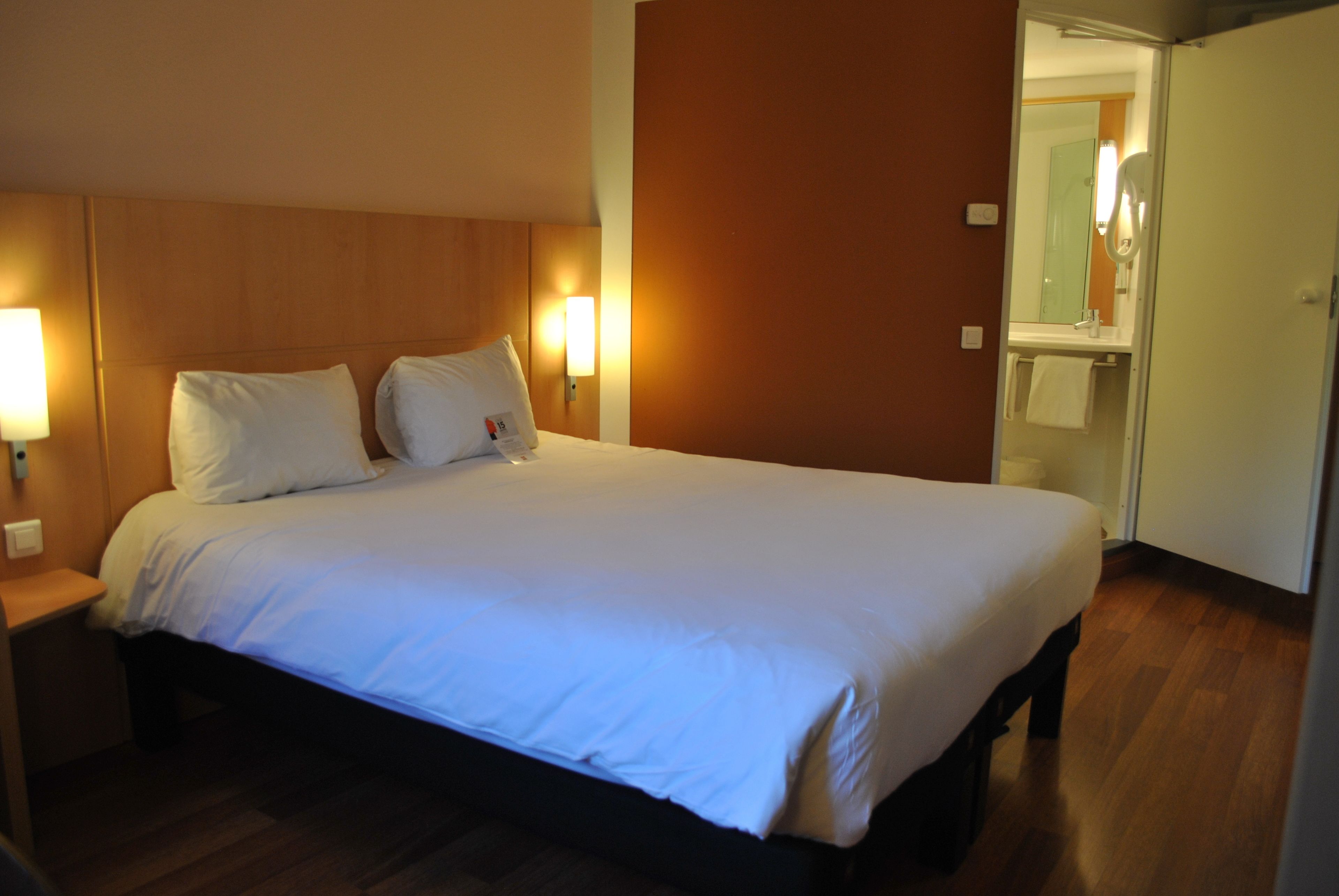 Un lit bien fait, avec du linge de lit blanc, dans une chambre d’hôtel : on aperçoit une tête de lit en bois, des lampes de chevet et une salle de bain à travers une porte ouverte.