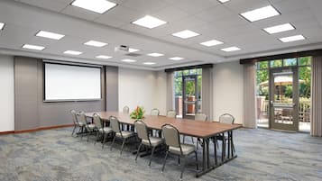 Un salón de conferencias con una larga mesa, sillas, una pantalla de proyector y ventanas grandes con cortinas.