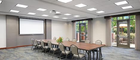 Un salón de conferencias con una larga mesa, sillas, una pantalla de proyector y ventanas grandes con cortinas.