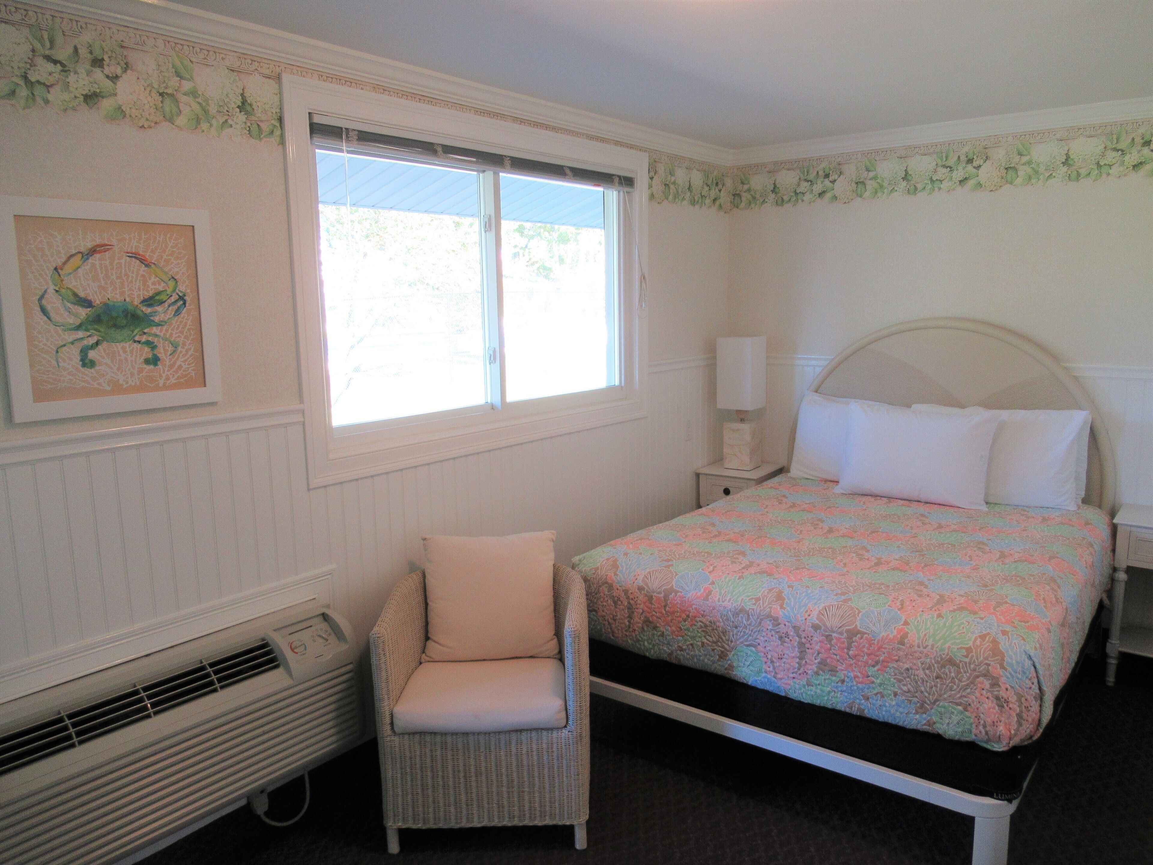 A bedroom with a bed, a chair, a nightstand, a window, and a wall-mounted heater.