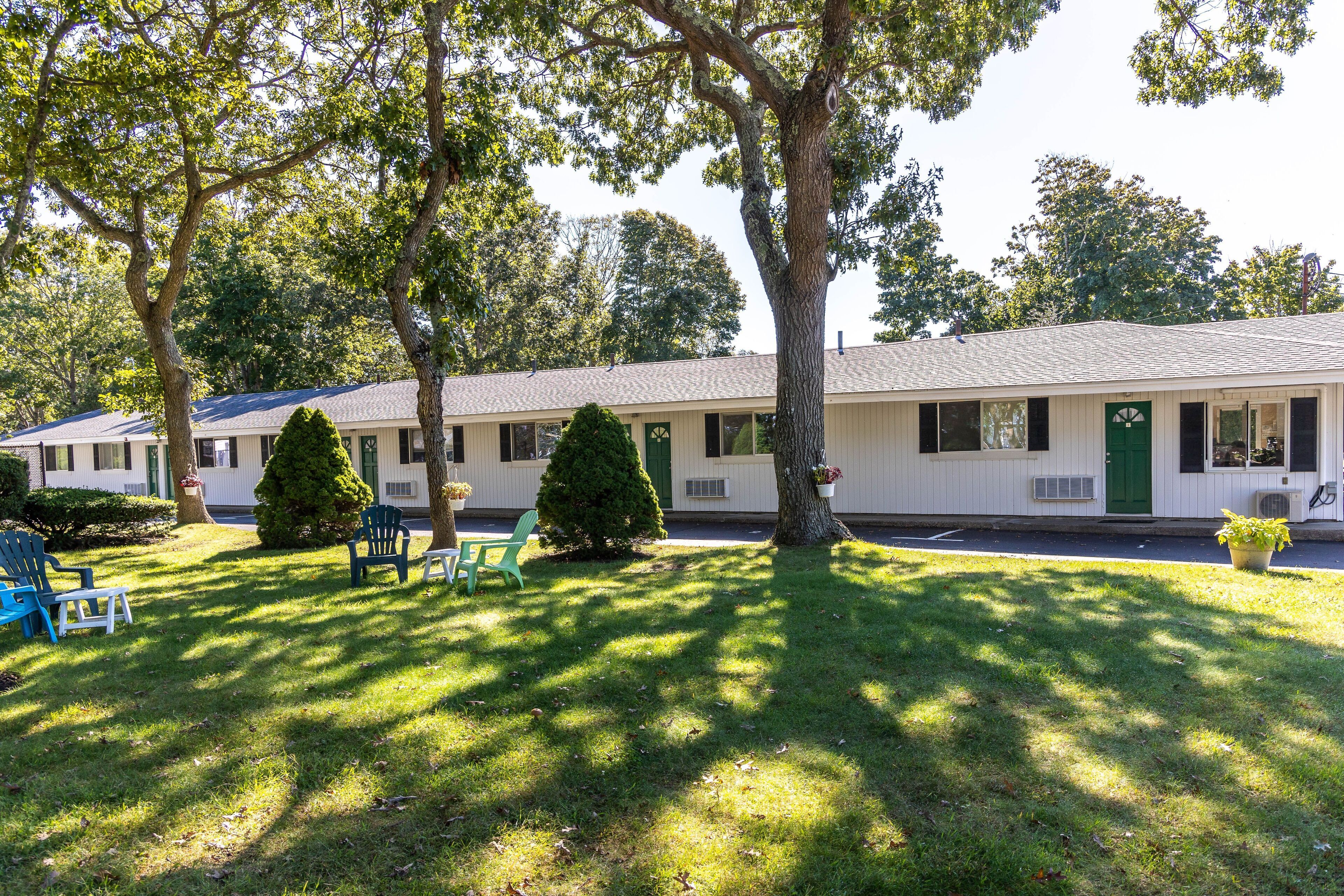 A motel with a row of rooms, each with a window and a door. There are trees and a lawn in front of the motel.