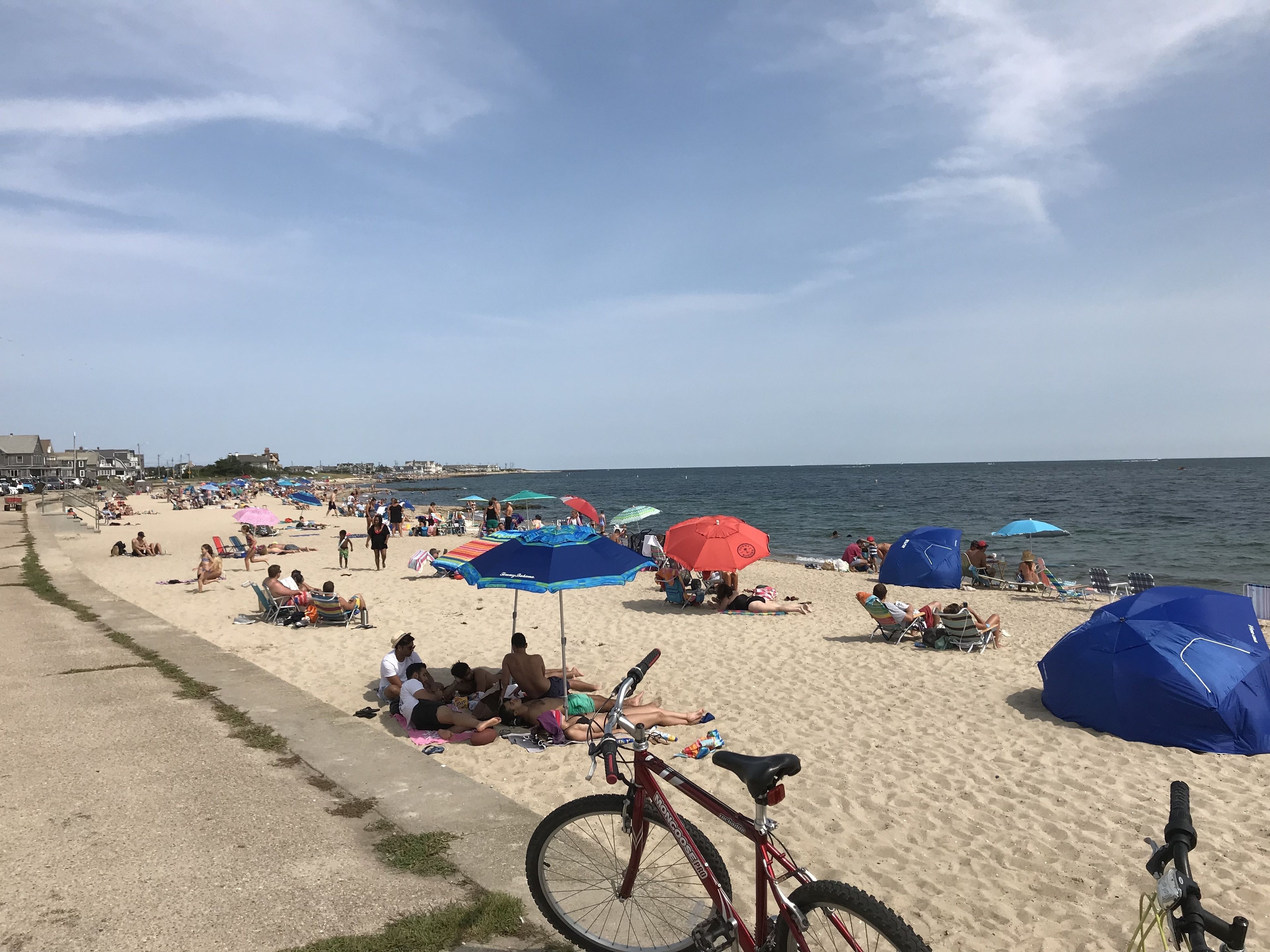 Una platja amb gent relaxant-se sota ombrel·les, una bicicleta i una vista clara de l'oceà.