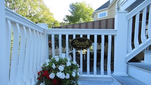 A wooden deck with a white railing, a potted plant with white and red flowers, and a sign that reads "Beach House".