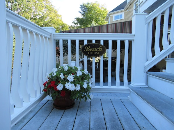 Una terraza de madera con barandilla blanca, una maceta con flores blancas y rojas, y un letrero que dice 'Beach House'.