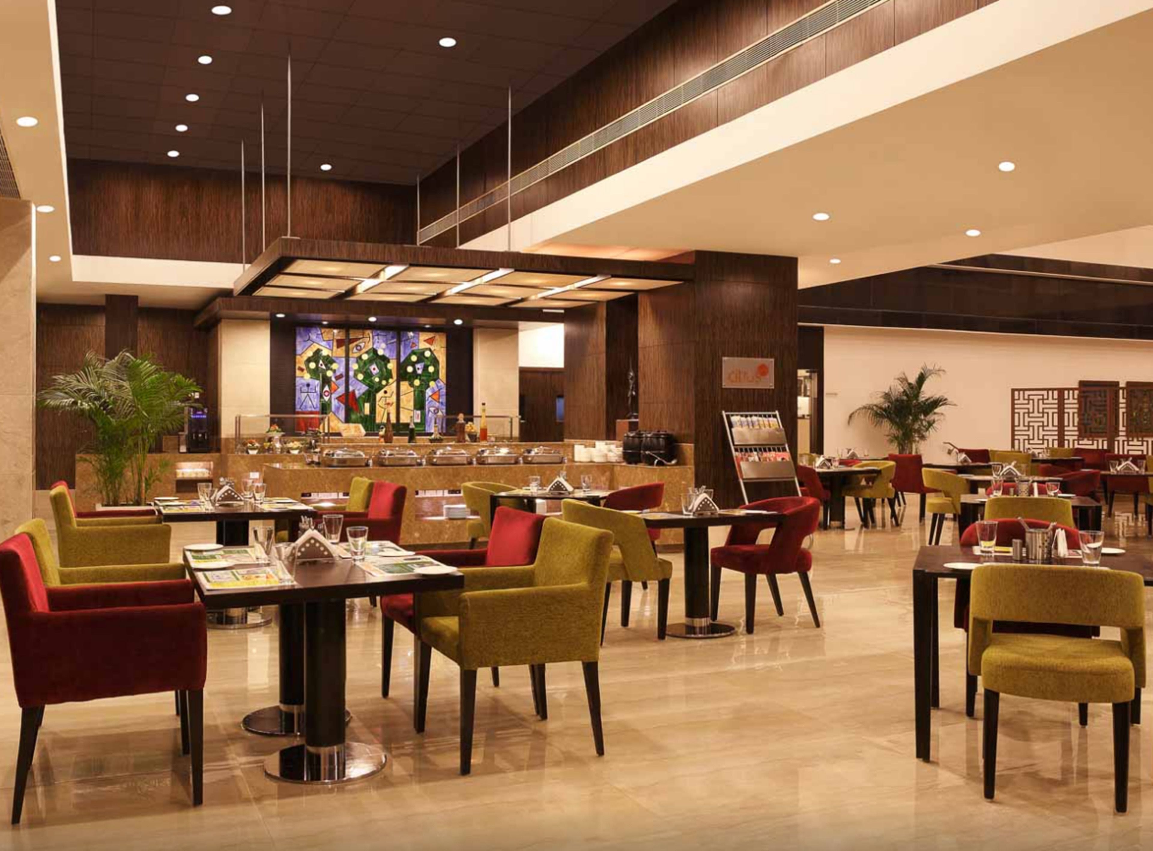 A modern hotel lobby with a central bar area, multiple dining tables with chairs, and a large decorative wall with a stained glass design.