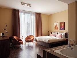 A modern hotel room with a bed, a desk, a chair, a large window with curtains, a sink, and two wall-mounted artworks.