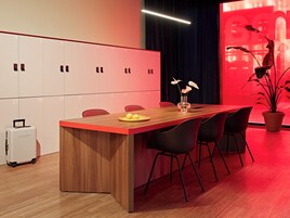 Ein moderner Konferenzraum mit einem Holztisch, schwarzen Stühlen, einer Topfpflanze und einer roten Akzentwand.