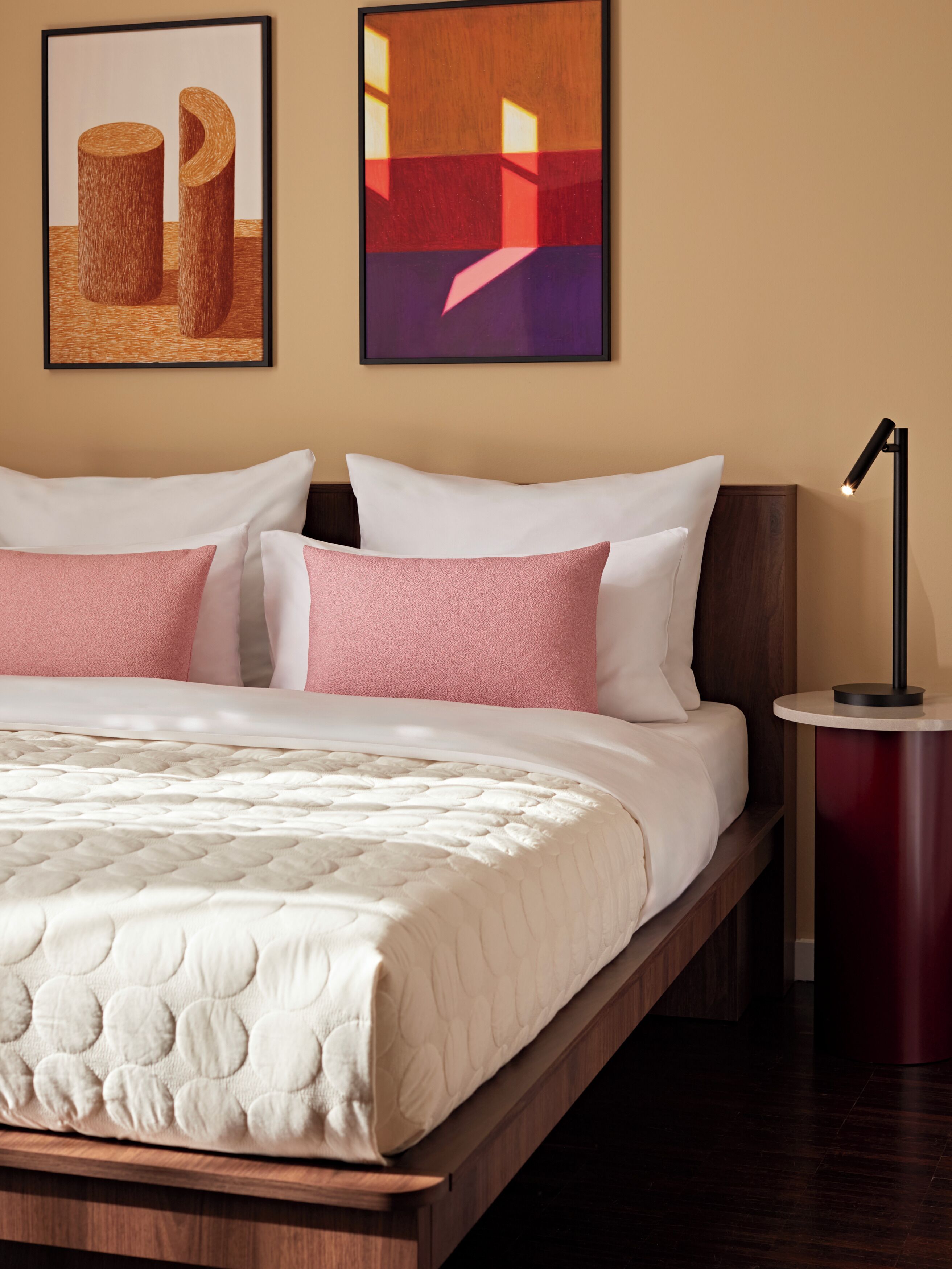 A neatly made bed with a white and pink color scheme, a bedside table with a lamp, and two abstract paintings on the wall.