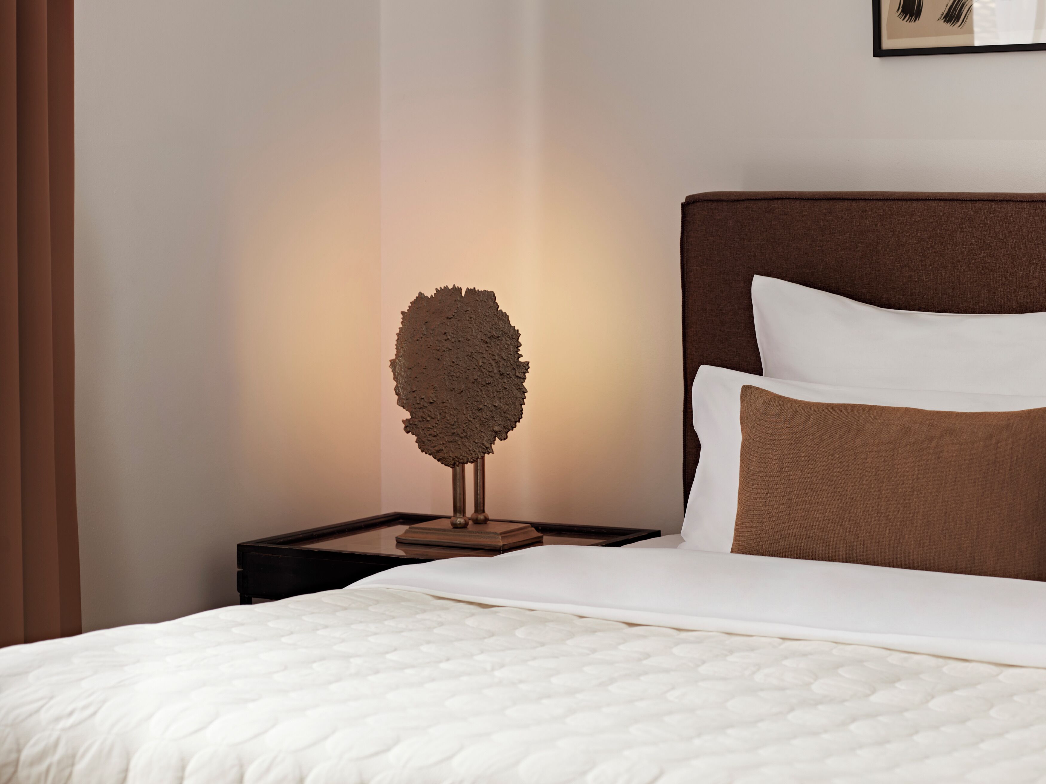 A neatly made bed with a white and brown pillow, a textured wall art piece, and a small wooden nightstand.