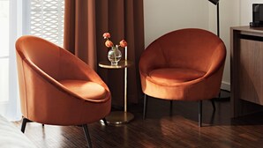 Deux fauteuils orange, une petite table ronde ornée d’un vase de fleurs, une lampe sur pied et un bureau avec un haut-parleur.