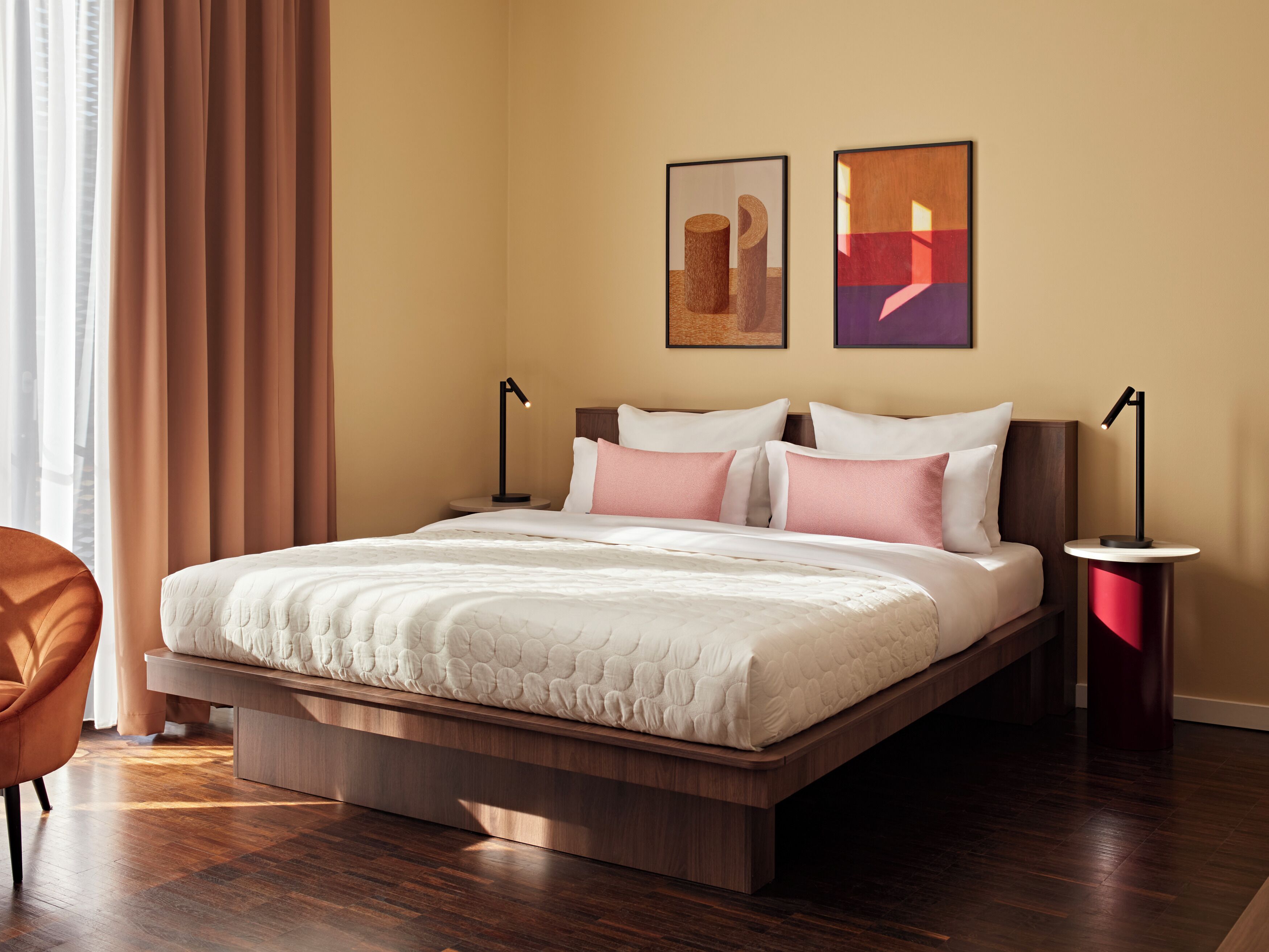 Una habitación de hotel moderna con un marco de cama de madera, una mesita de noche, una silla y dos cuadros enmarcados en la pared.