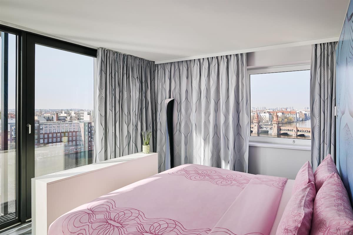 A bedroom with a large window, a bed with pink bedding, and a view of a cityscape.