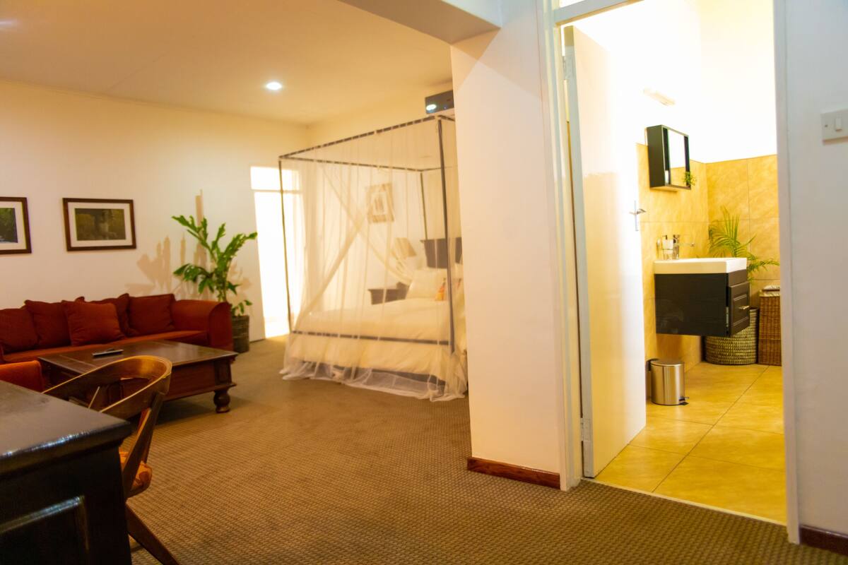 Habitación de hotel con cama, sofá, una mesita de centro, una planta y un baño visible a través de una puerta abierta.