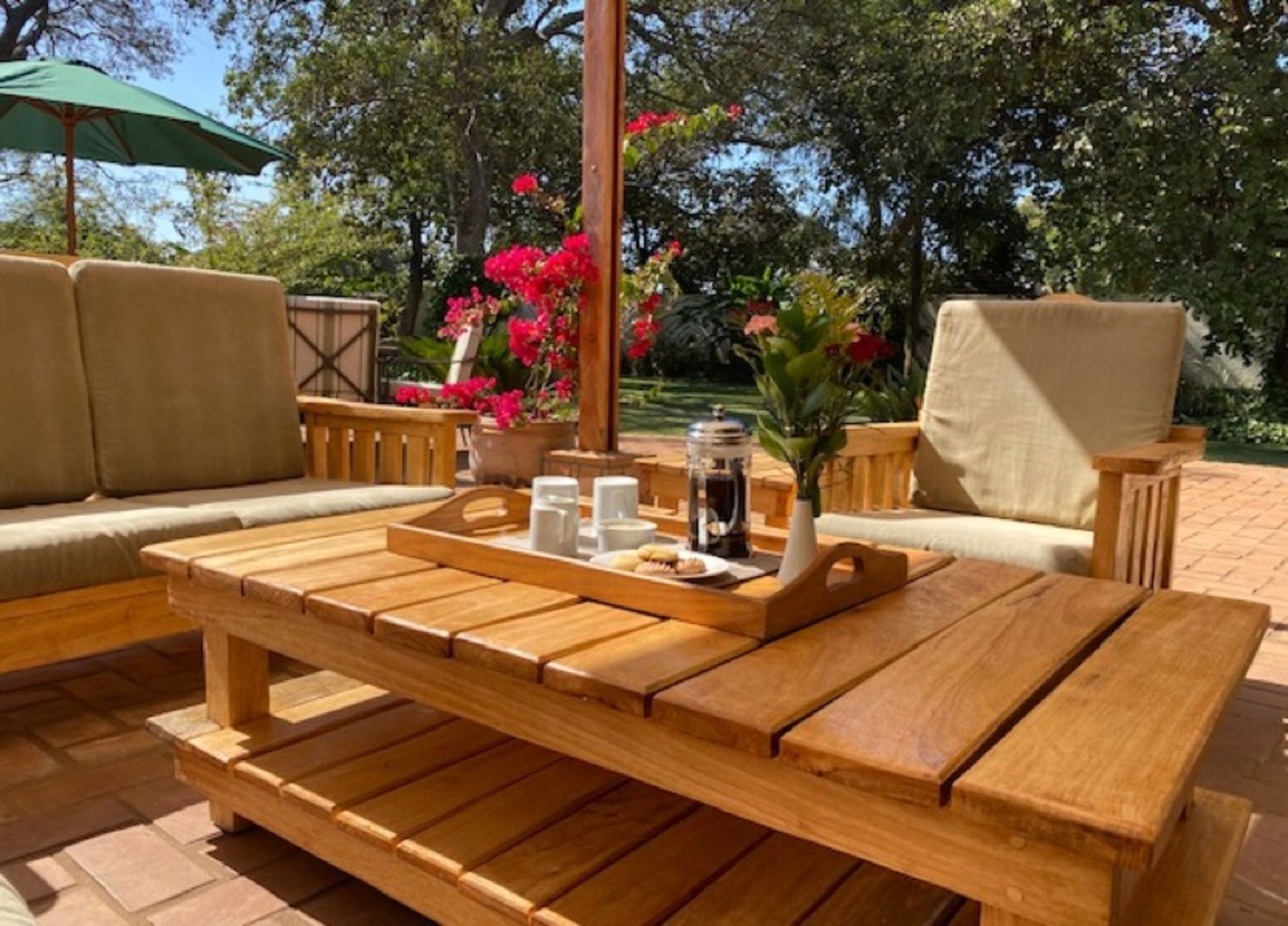 Un área de estar al aire libre con muebles de madera, una bandeja con alimentos para el desayuno, como pasteles y café, rodeada de abundante follaje y una cerca de madera.
