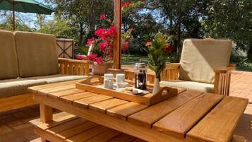Un área de estar al aire libre con muebles de madera, una bandeja con alimentos para el desayuno, como pasteles y café, rodeada de abundante follaje y una cerca de madera.