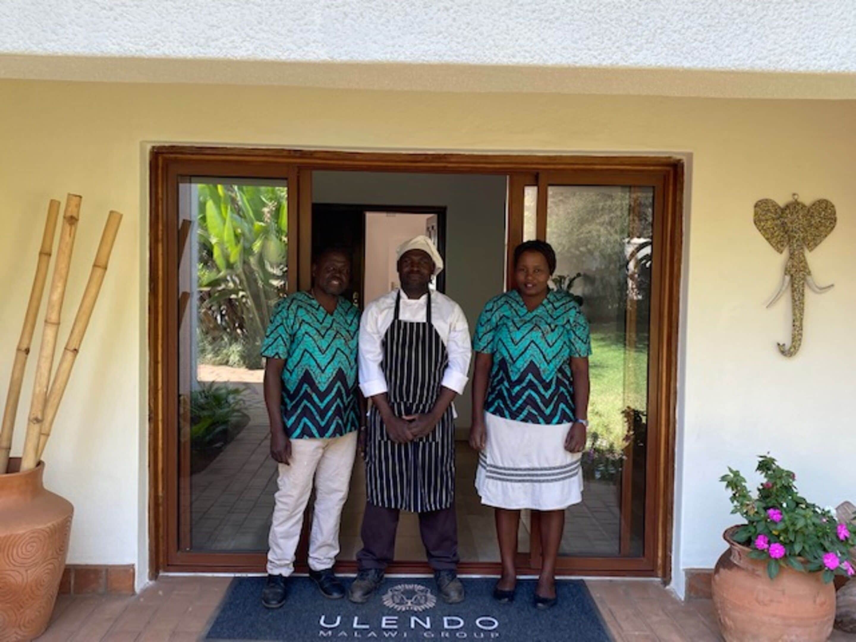 Trois personnes se tiennent devant l’entrée d’un bâtiment, devant un paillasson sur lequel est inscrit « ULENDO MALAWI GROUP ».