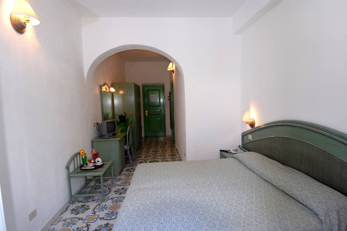 Une chambre d’hôtel étroite, avec un lit, un bureau sur lequel se trouve une télévision et une porte verte.