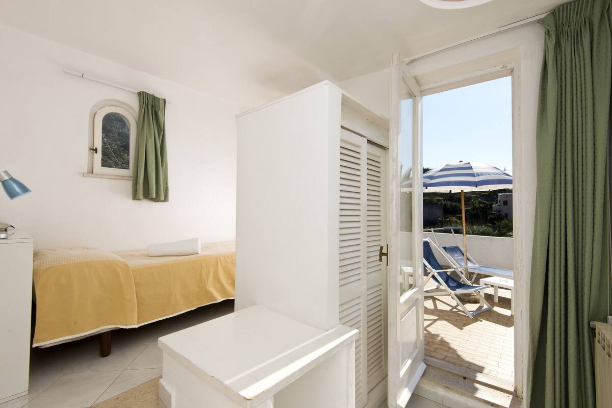 Une chambre avec un lit, une petite table et une armoire. Il y a une fenêtre avec des rideaux et une vue sur un espace extérieur agrémenté d’un parasol.