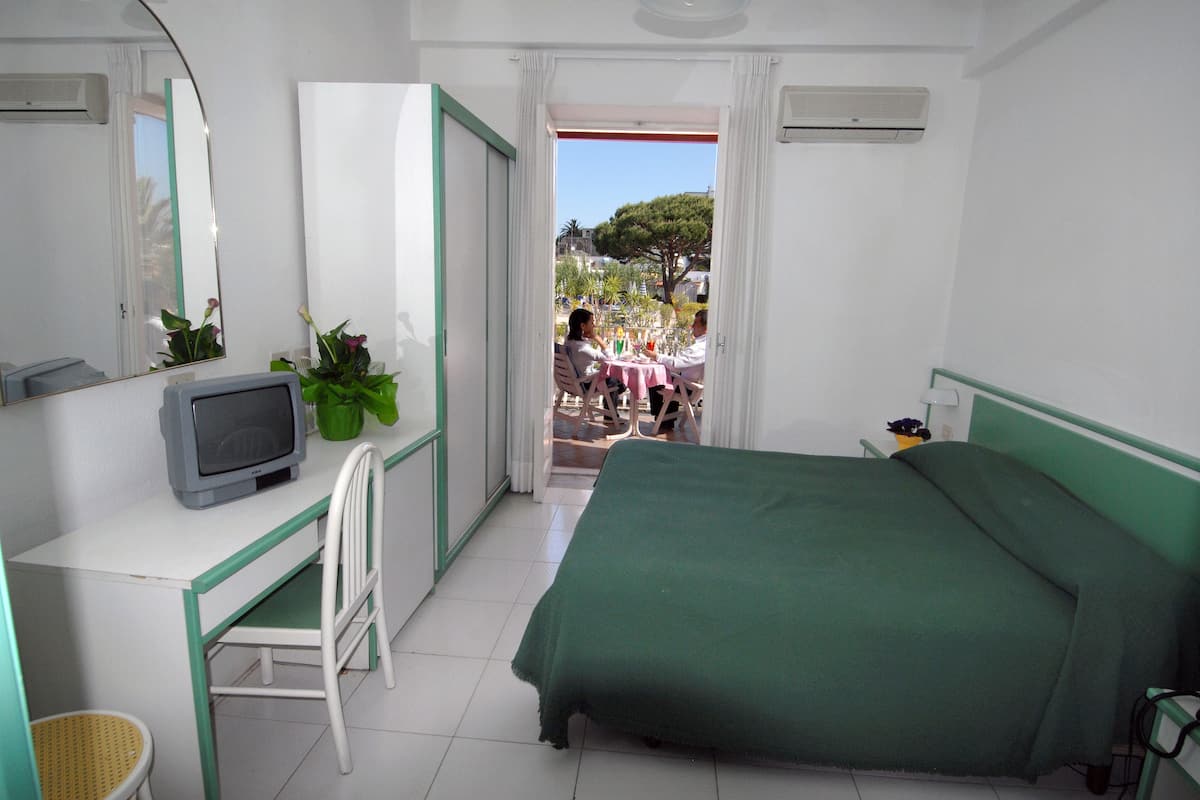 Une chambre avec un lit, un bureau avec une télévision, une chaise et une vue sur un espace extérieur où se trouvent des personnes et des plantes.
