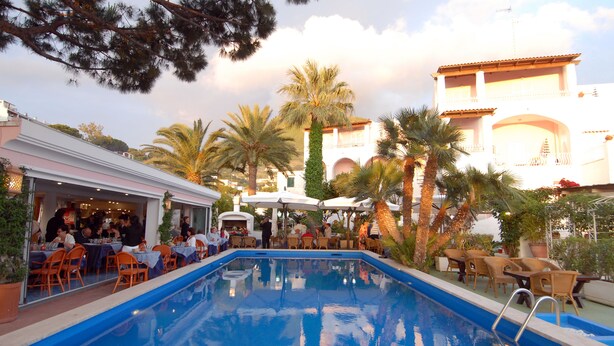 A poolside area with tables and chairs, a pool, and palm trees. - Hotel Terme Colella (Forio)