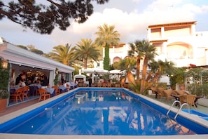 Un área junto a la piscina con mesas y sillas, piscina y palmeras.