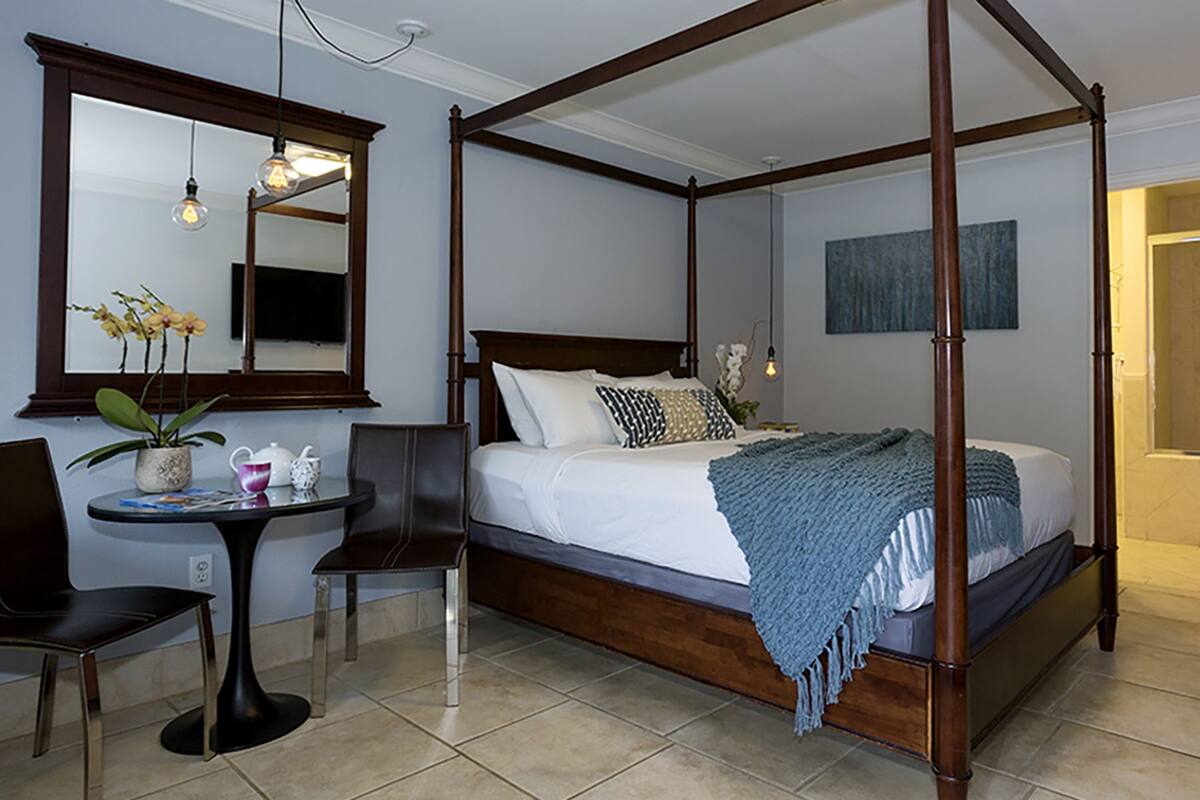 A four-poster bed with a wooden frame, a bedside table with a lamp, a chair, and a small table with a plant and a teapot.