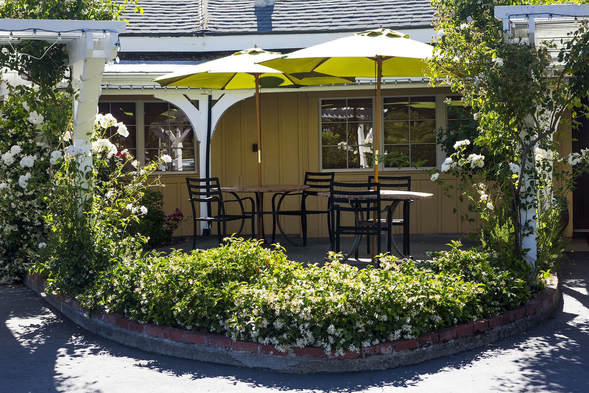 Un área de estar al aire libre con una mesa y sillas bajo un paraguas amarillo, rodeada por una jardinera de ladrillo con flores blancas.
