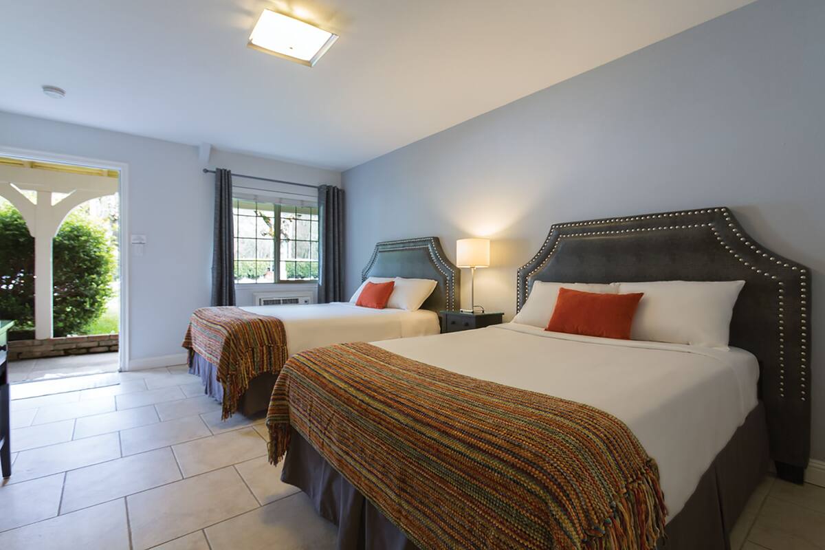 A hotel room with two beds, a window with curtains, a lamp, and a view of greenery outside.