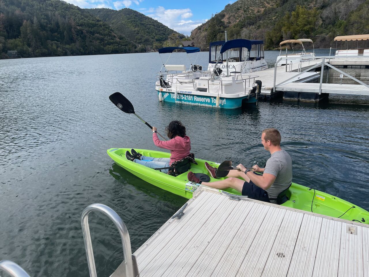 A man and a woman kayaking on a green kayak on a calm lake with a dock and a boat in the background.