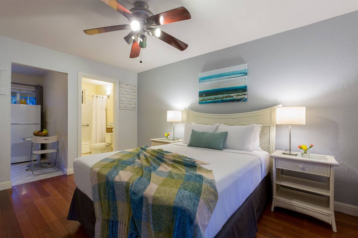 A hotel room with a bed, two bedside tables with lamps, a ceiling fan, a painting on the wall, and a view of the bathroom.