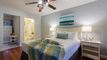 Habitación de hotel con cama, dos mesitas de noche con lámparas, ventilador de techo, un cuadro en la pared y vista al baño.
