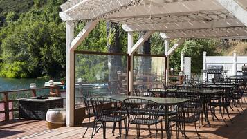 An outdoor dining area with tables and chairs, overlooking a body of water and surrounded by greenery.