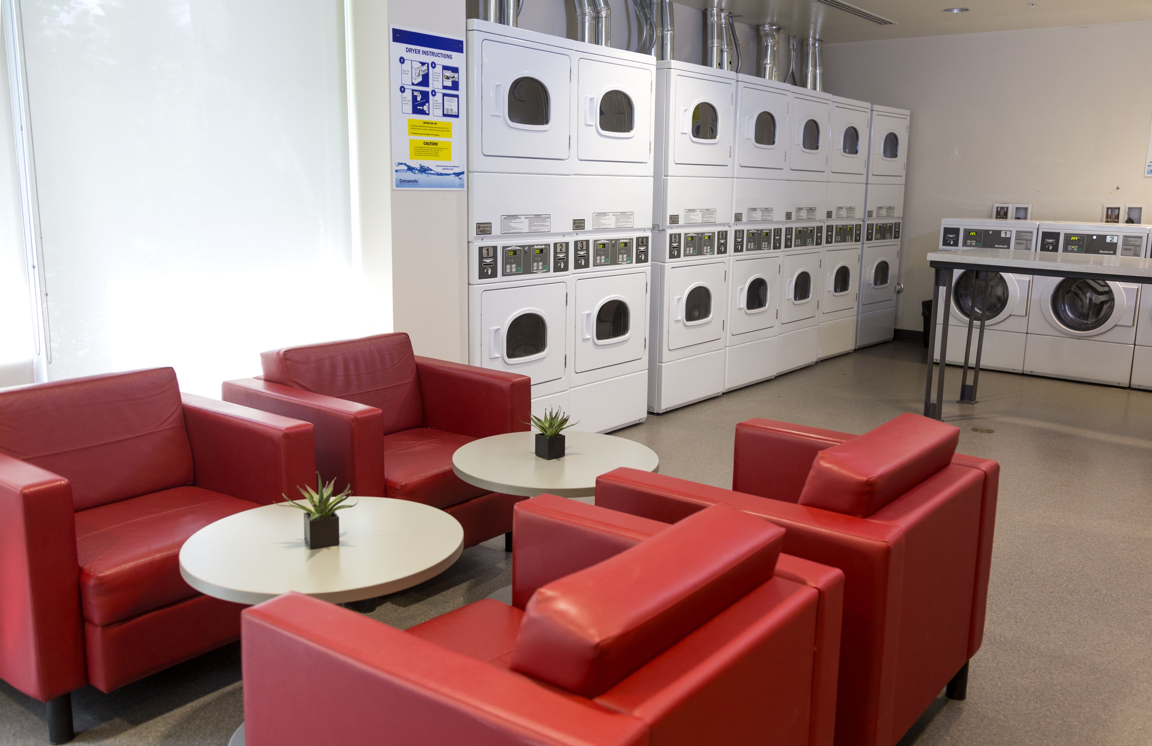 A laundry facility with multiple washing machines and dryers, red seating area, and a small table with a plant.