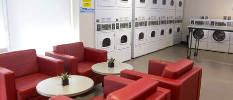 A laundry facility with multiple washing machines and dryers, red seating area, and a small table with a plant.