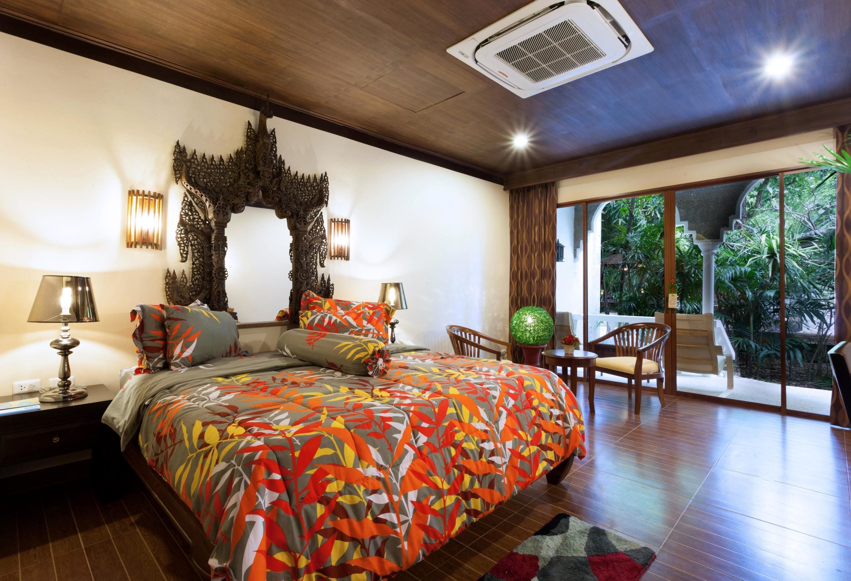 A bedroom with a large bed, a nightstand, a lamp, a decorative wall piece, and a view of a lush garden through a sliding door.
