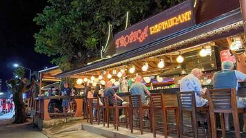 Um restaurante ao ar livre com cadeiras e mesas de madeira, um letreiro de neon e uma árvore ao fundo.
