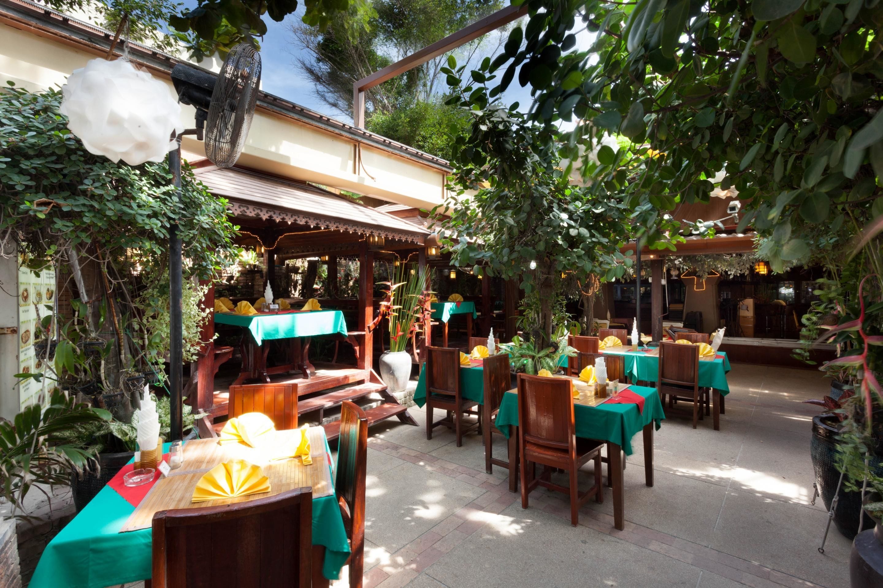 An outdoor dining area with tables covered in green tablecloths and wooden chairs, surrounded by lush greenery and a traditional architectural style.
