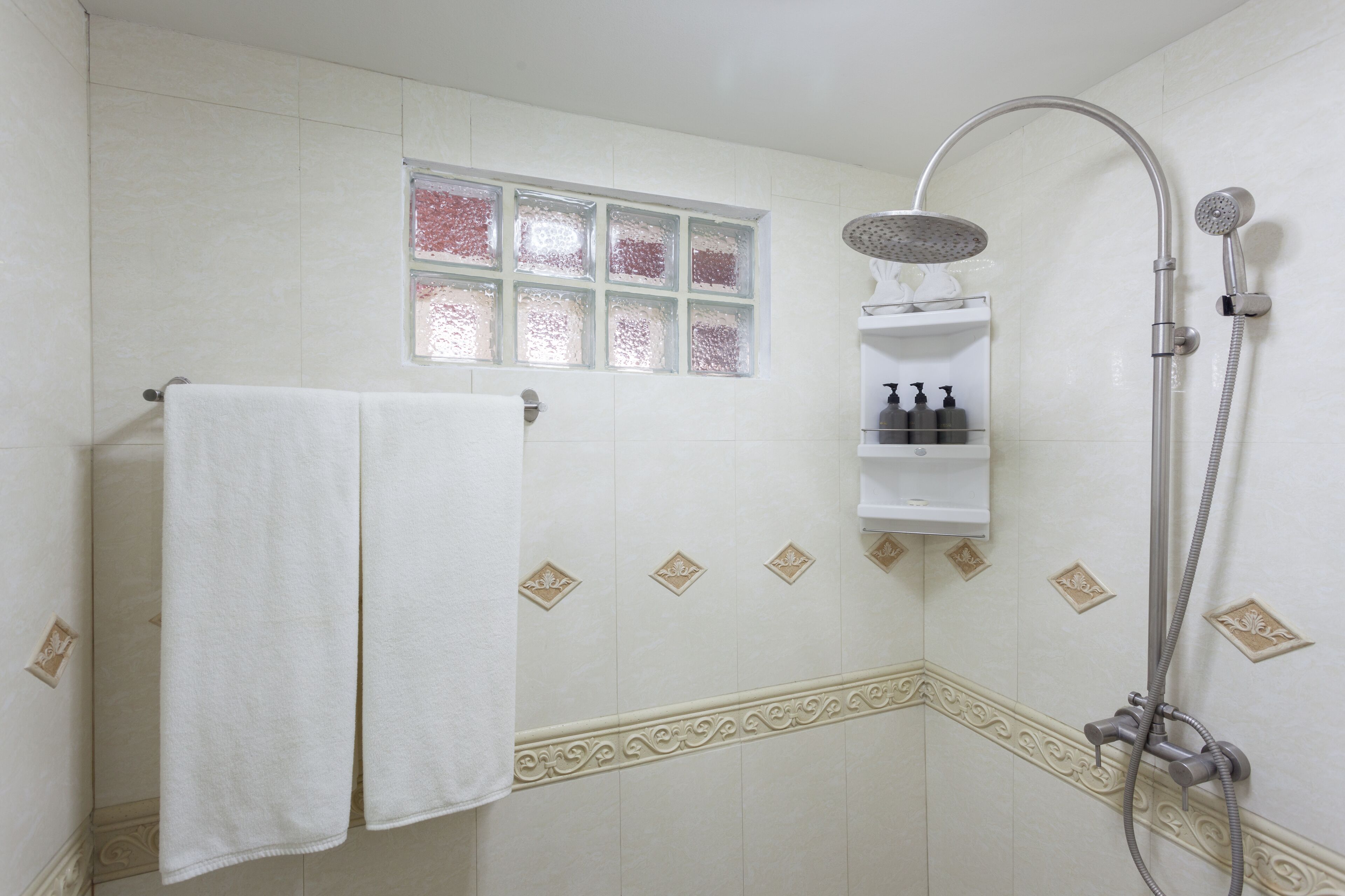 A bathroom with a shower, a towel rack, and a window with decorative tiles.