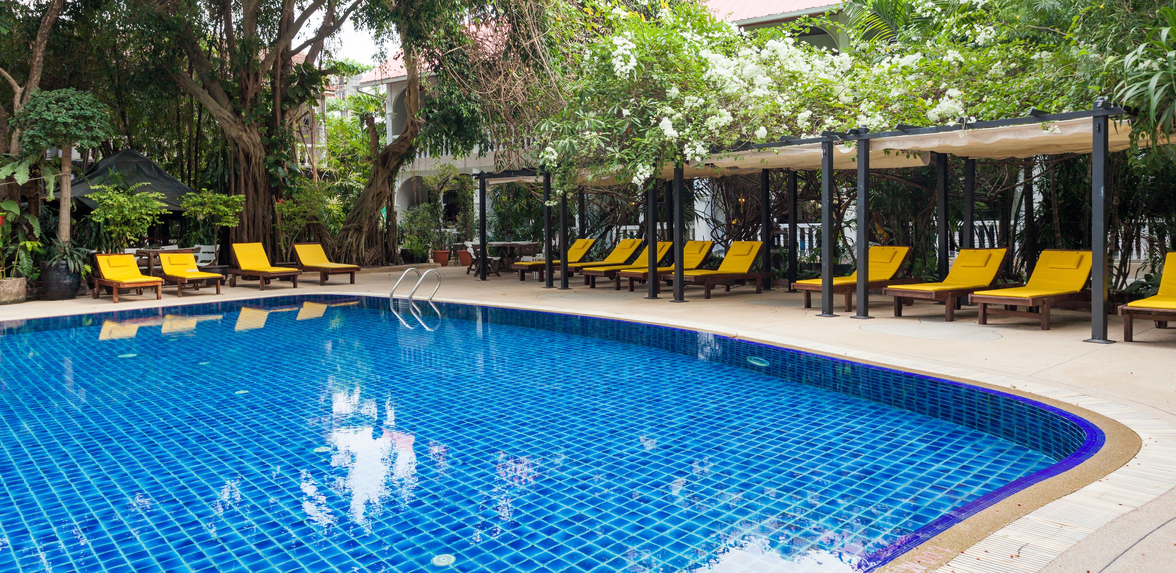 A swimming pool with tiled blue floor, surrounded by yellow lounge chairs and covered seating areas, with trees and greenery in the background.