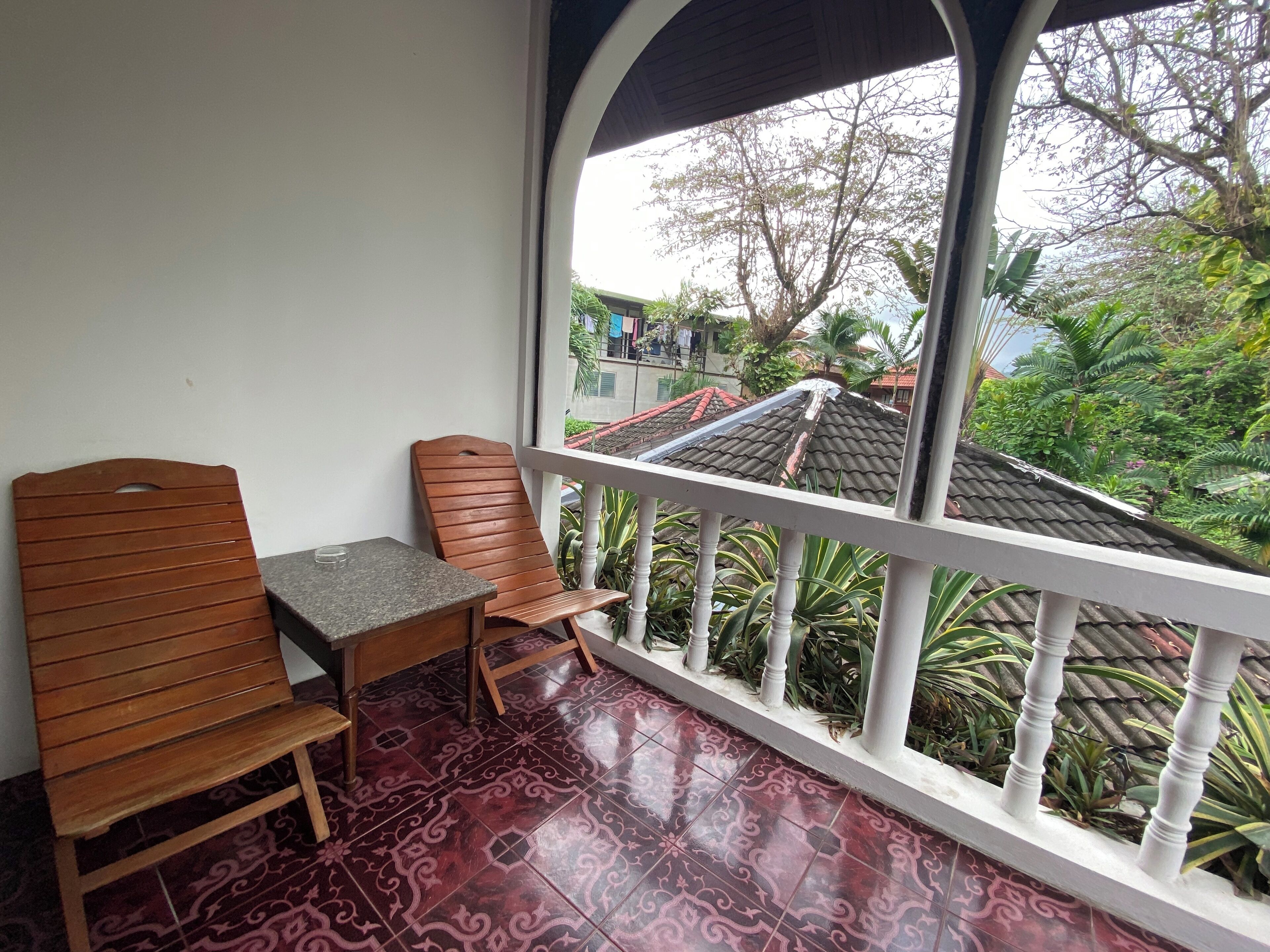 A balcony with a wooden chair and table, a patterned floor, and a view of a residential area with trees and a tiled roof.