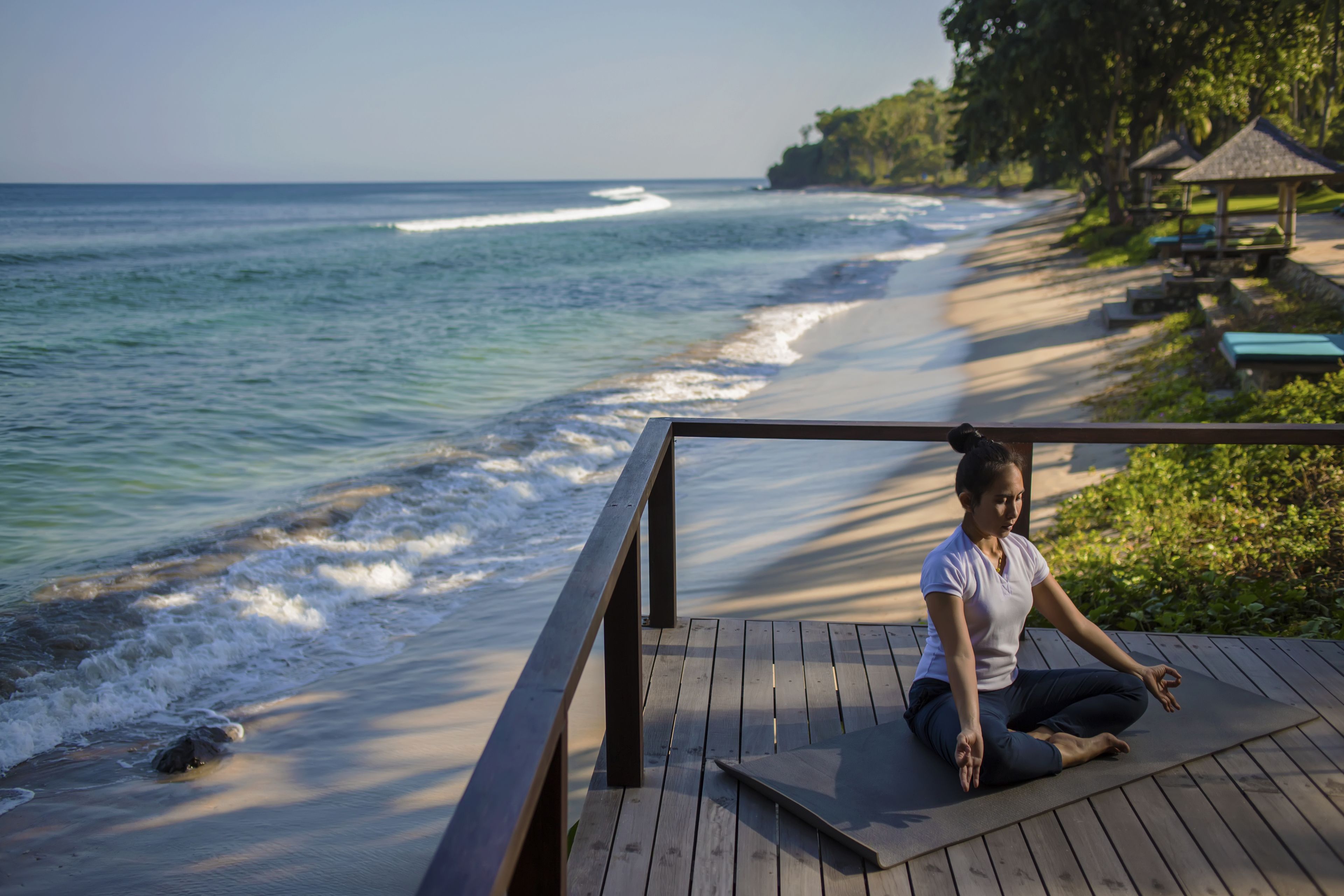 A person meditating on a wooden deck overlooking a beach with waves, a gazebo, and lounge chairs.