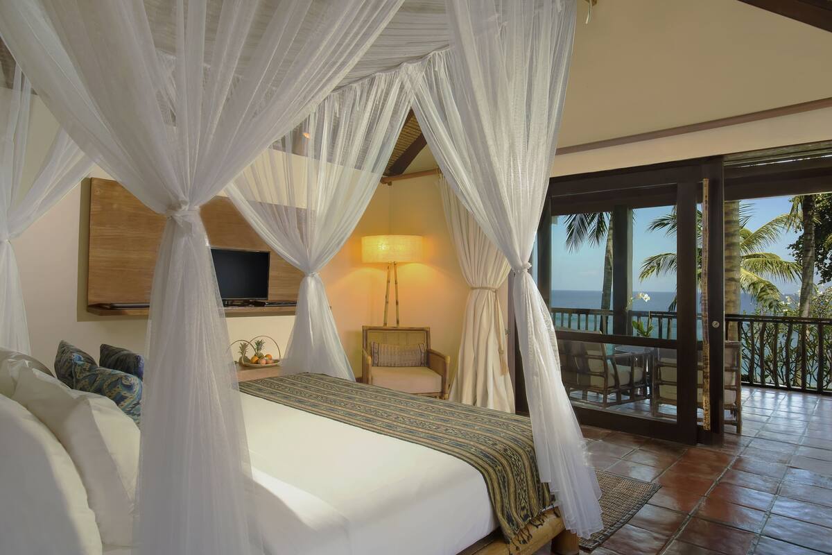 A bedroom with a bed, a TV, a chair, and a balcony with a view of palm trees.