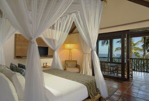 Un dormitorio con cama, televisor, sillón y balcón con vista a palmeras.