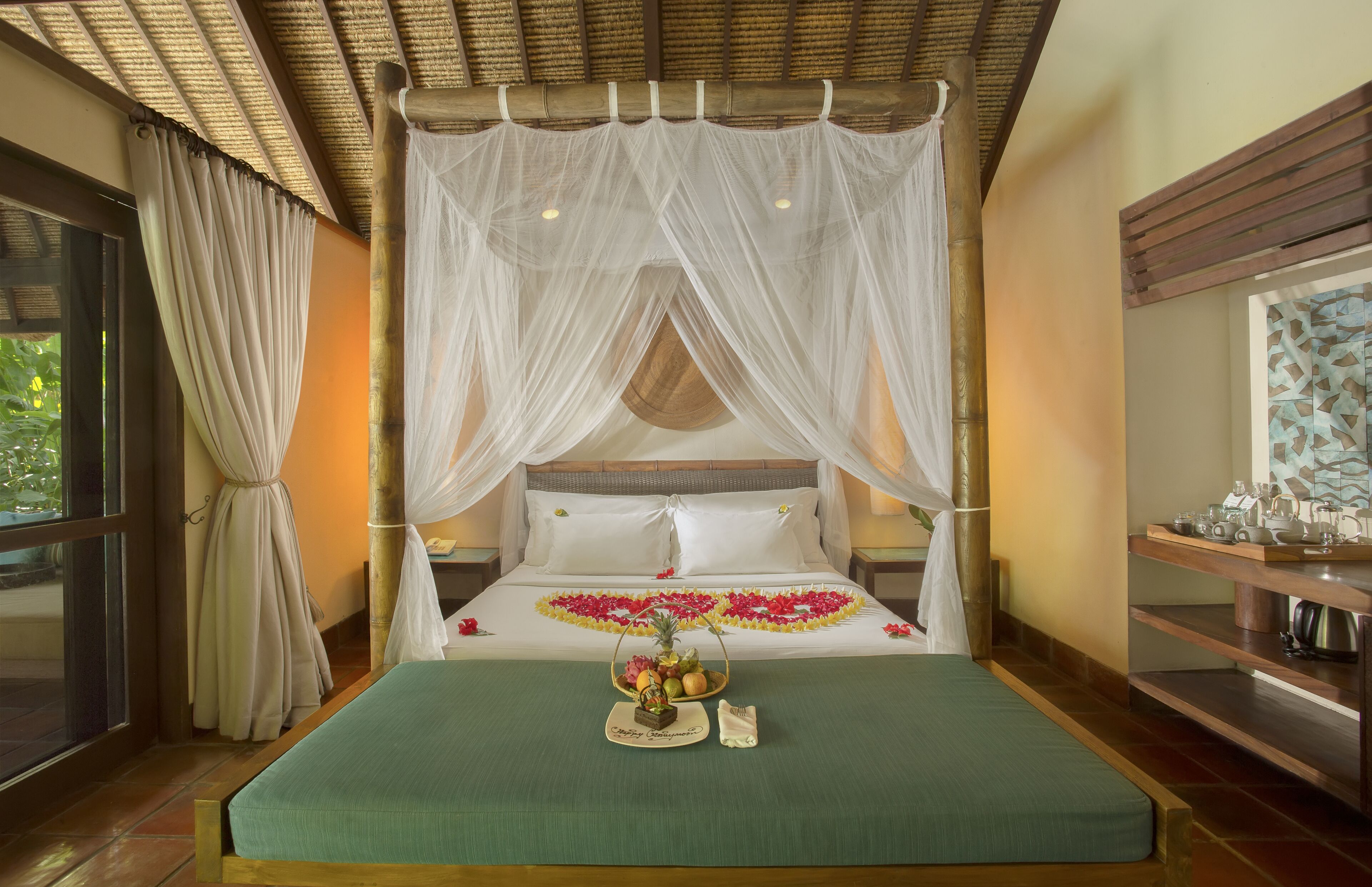 A four-poster bed with a canopy, a bedside table with a tray of fruit and flowers, and a view of the outdoors through a large window.